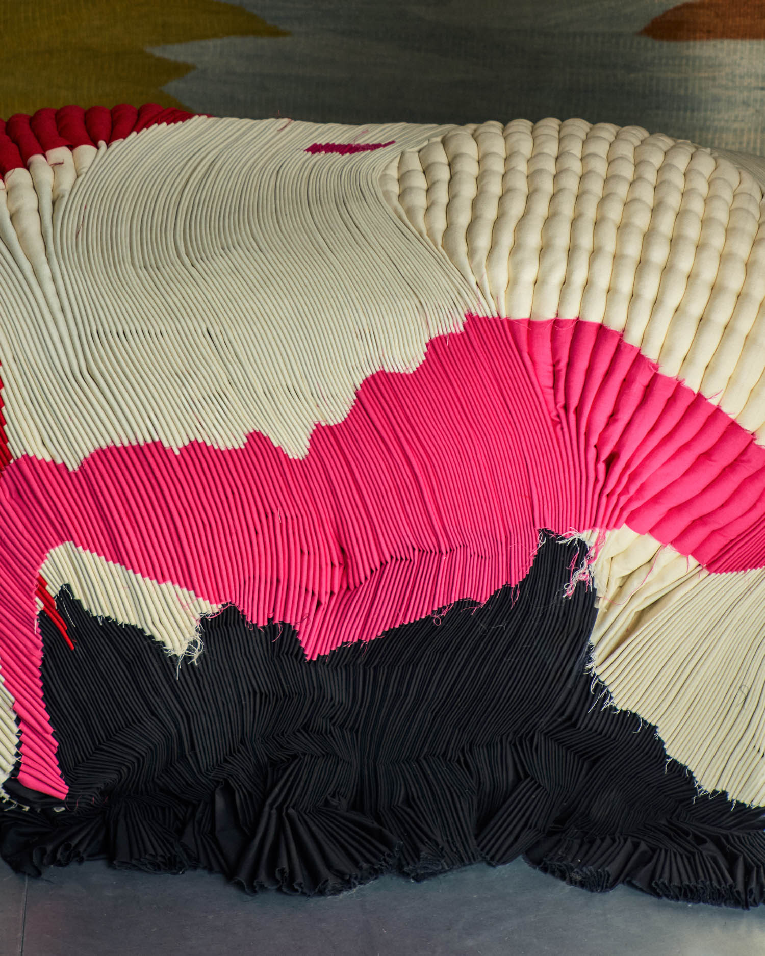 A textured fabric sculpture with pleated surfaces in cream, pink, and black sits on a smooth floor, with a multicolored carpet partially visible in the background.