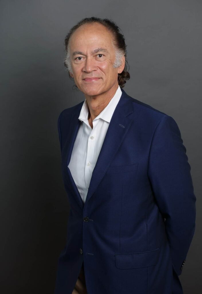 A man wearing a dark blue suit and white dress shirt stands in front of a plain gray background, looking at the camera with a slight smile and his hands behind his back.