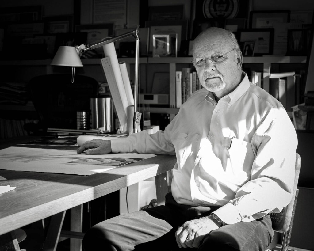 An older man with glasses sits at a desk covered with papers and books in an office, looking at the camera. The room is filled with shelves, framed certificates, and a desk lamp.