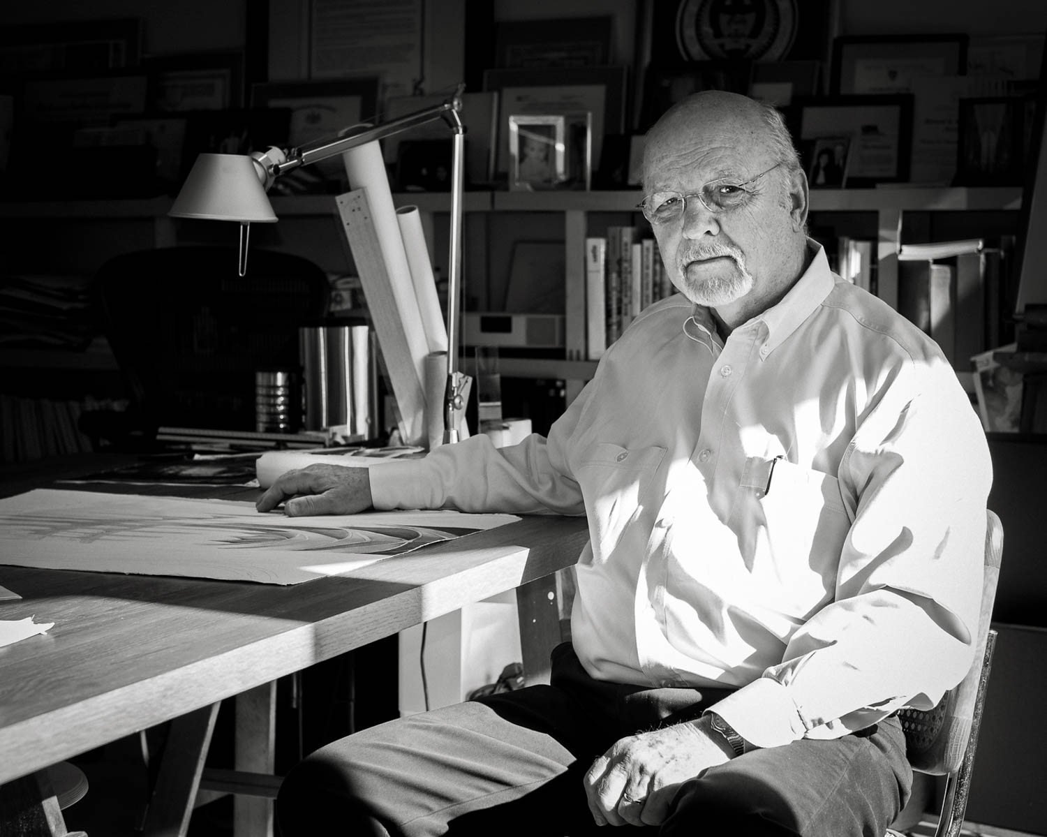 An older man with glasses sits at a desk covered with papers and books in an office, looking at the camera. The room is filled with shelves, framed certificates, and a desk lamp.