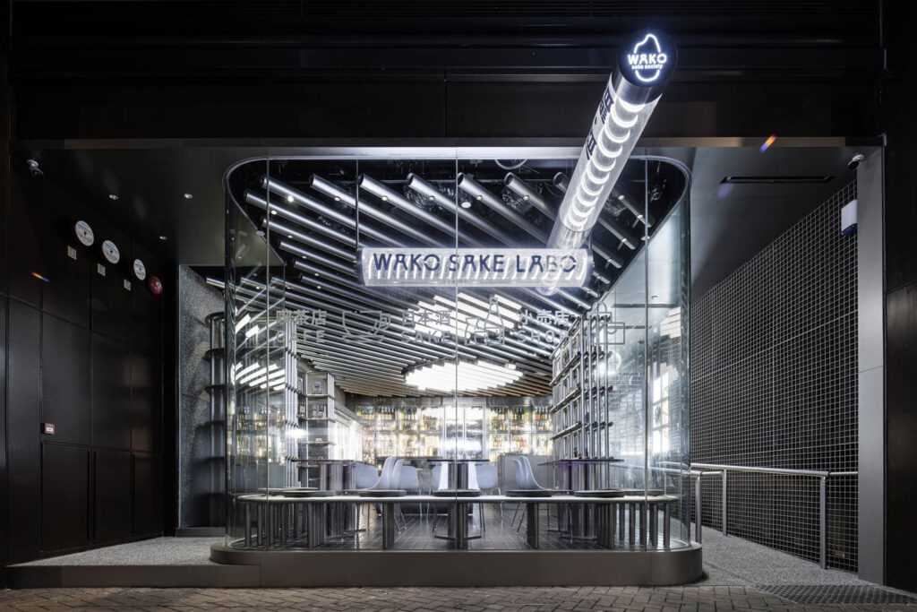 Modern restaurant exterior with glass walls, illuminated signage reading “WAKO SAKELABO,” metal tables, and a ceiling with linear LED lights.