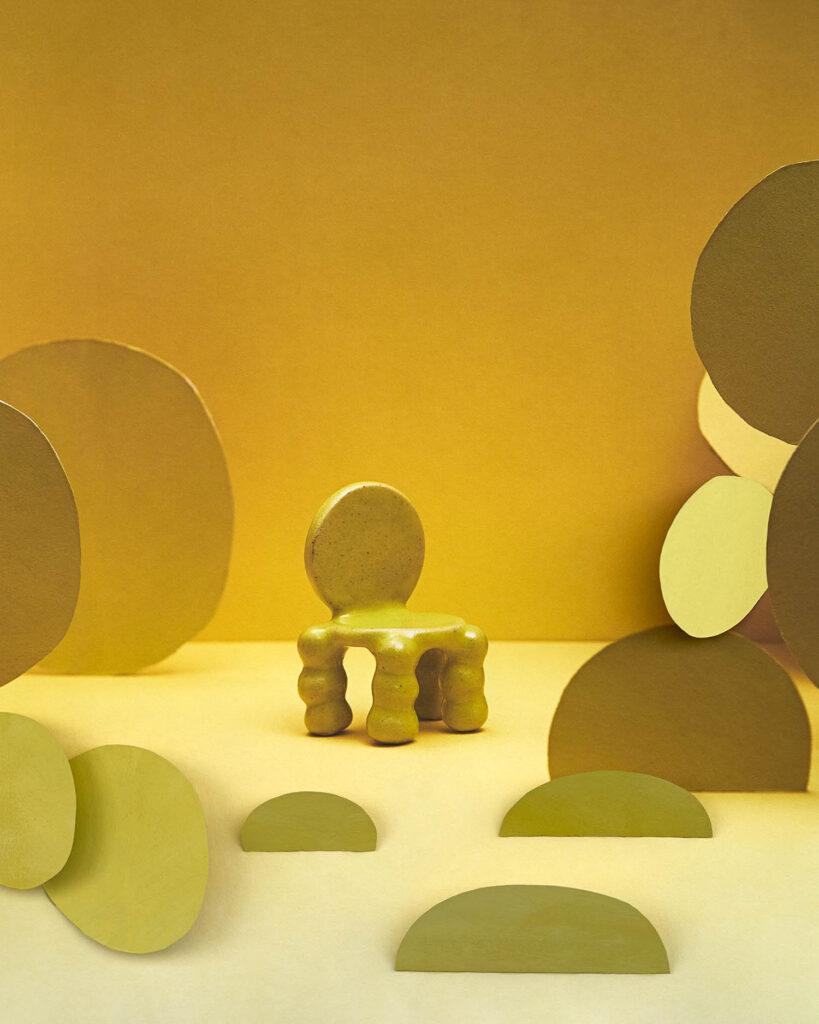 A small, round-backed chair with wavy legs sits on a yellow surface surrounded by flat, oval and semi-circle shapes against a yellow background.