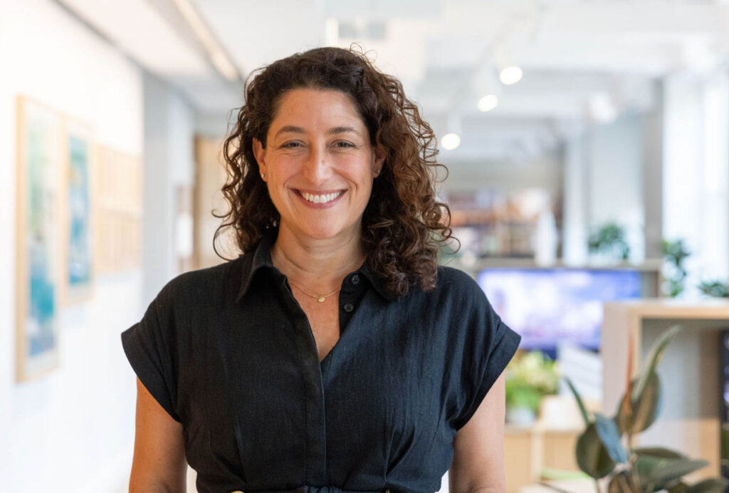 A person with curly brown hair wearing a dark short-sleeved shirt smiles while standing in a modern, brightly lit office space.