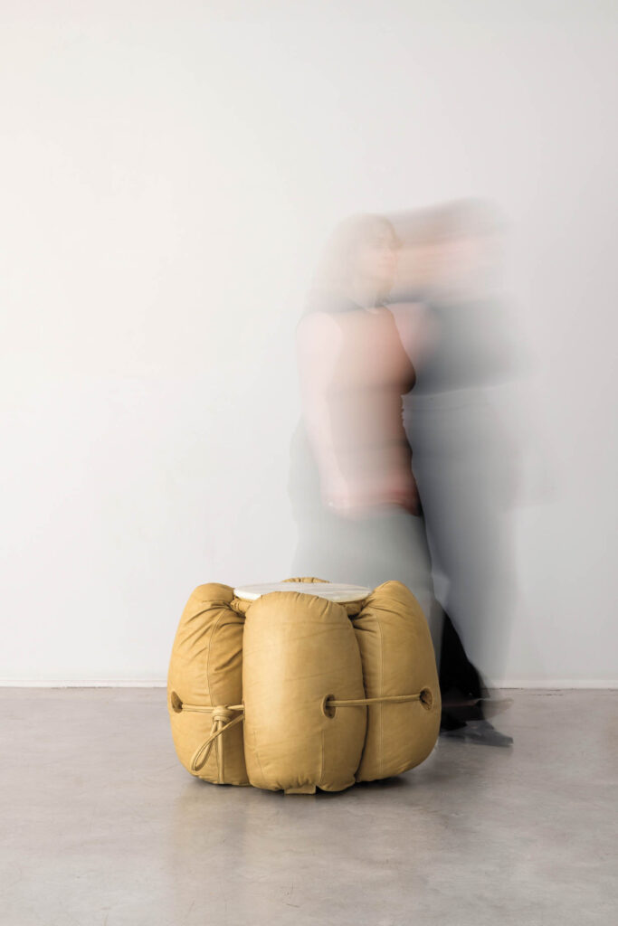A blurred person walks past a yellow, cushion-like object tied with cords on a gray floor against a plain white wall.