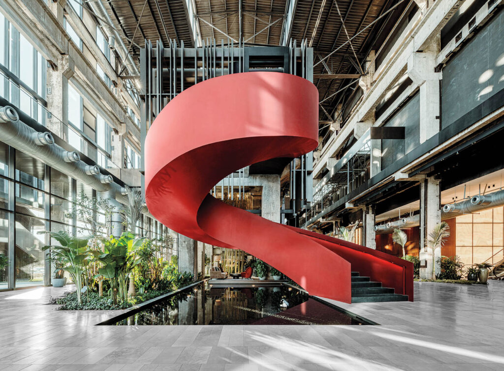 A large red spiral staircase stands over a reflective pool in an industrial-style building with exposed beams, large windows, and indoor plants.