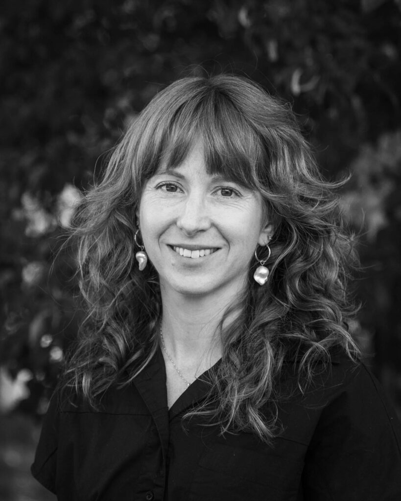 Woman with wavy hair wearing a dark shirt and large earrings, smiling at the camera in an outdoor setting with blurred foliage in the background.
