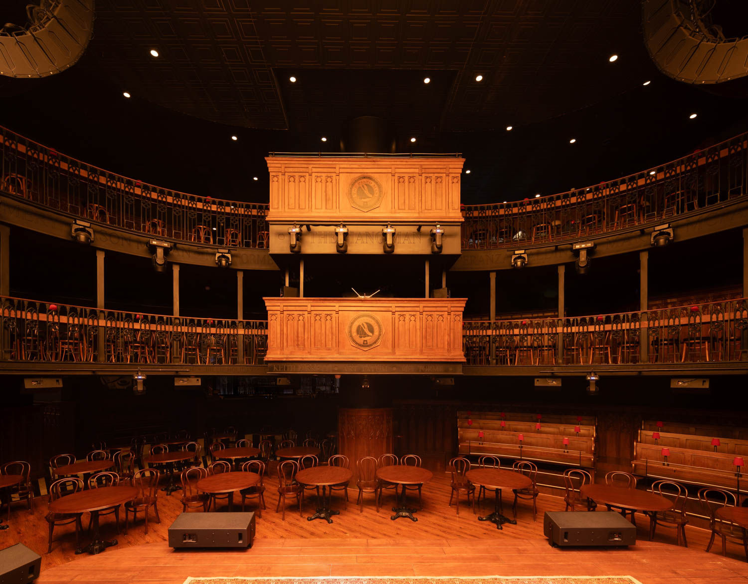Empty theater with round tables and wooden chairs on the main floor, two balcony levels with seating, and ornate wooden box seats in the center above the stage.