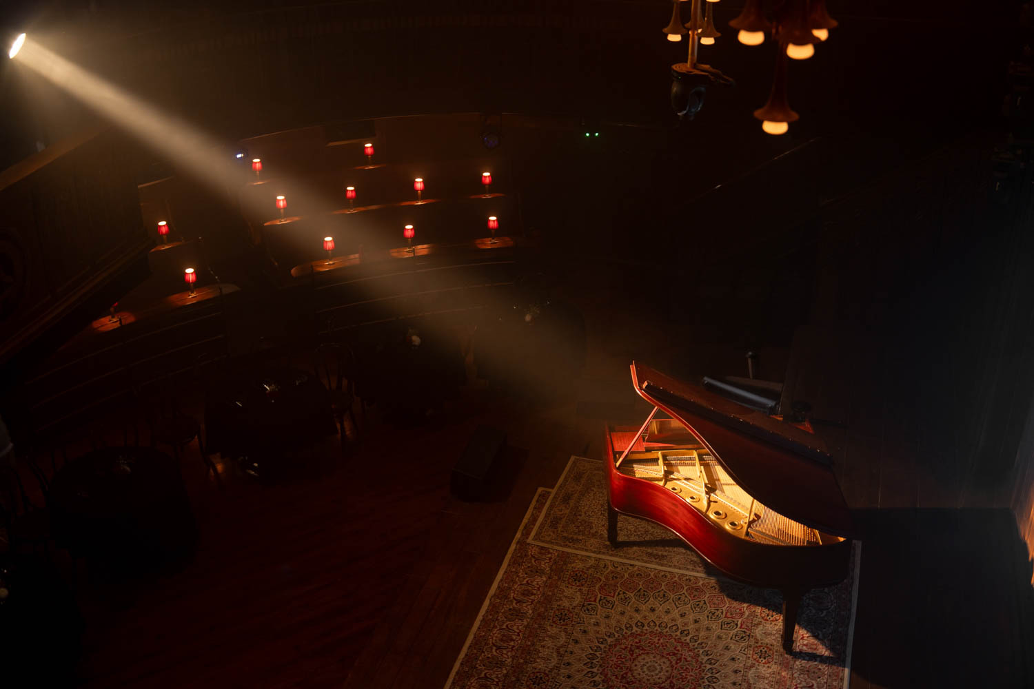 A spotlight shines on an open grand piano on a stage with a rug, while small red lights illuminate empty seats in a dark, intimate venue.