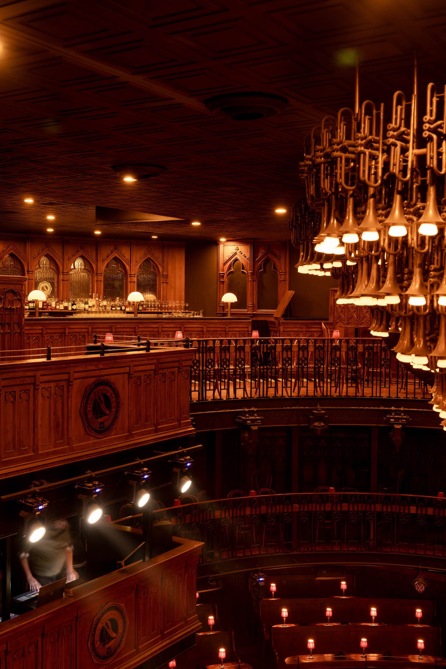Elegant, dimly lit theater interior with wooden balconies, ornate chandelier, and a bar in the background. Spotlights illuminate a person in a booth below.