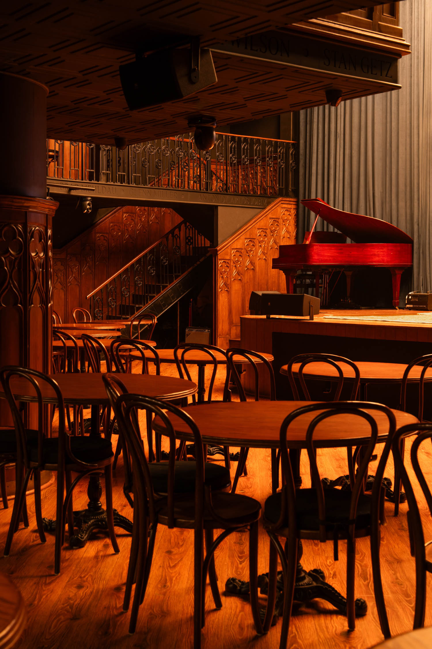 A dimly lit jazz club with wooden tables and chairs, a staircase, and a red grand piano on stage near blue curtains.