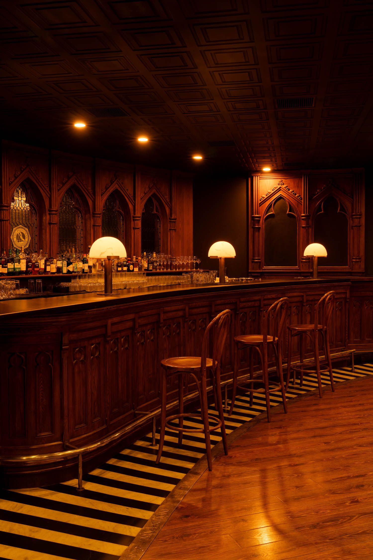 Dimly lit bar with ornate wooden paneling, arched mirrors, a row of wooden stools, and three round table lamps on the counter.