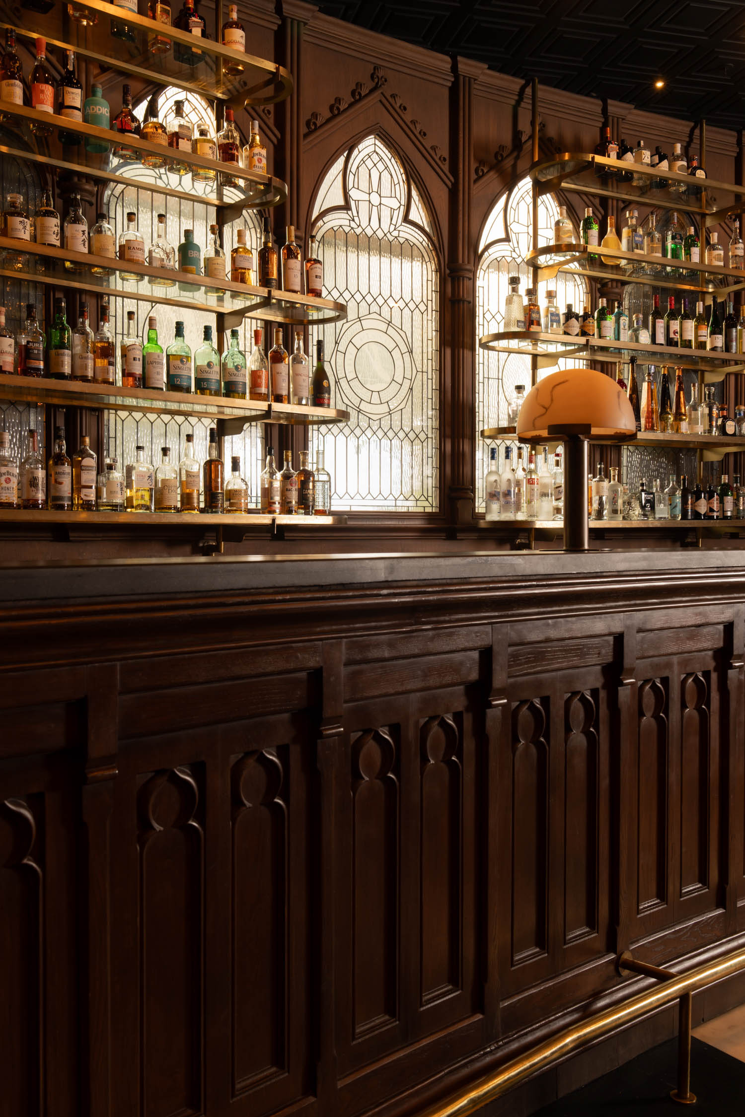 A wooden bar counter with ornate paneling faces shelves of liquor bottles and a stained glass window. Warm lighting and a retro lamp create a cozy atmosphere.
