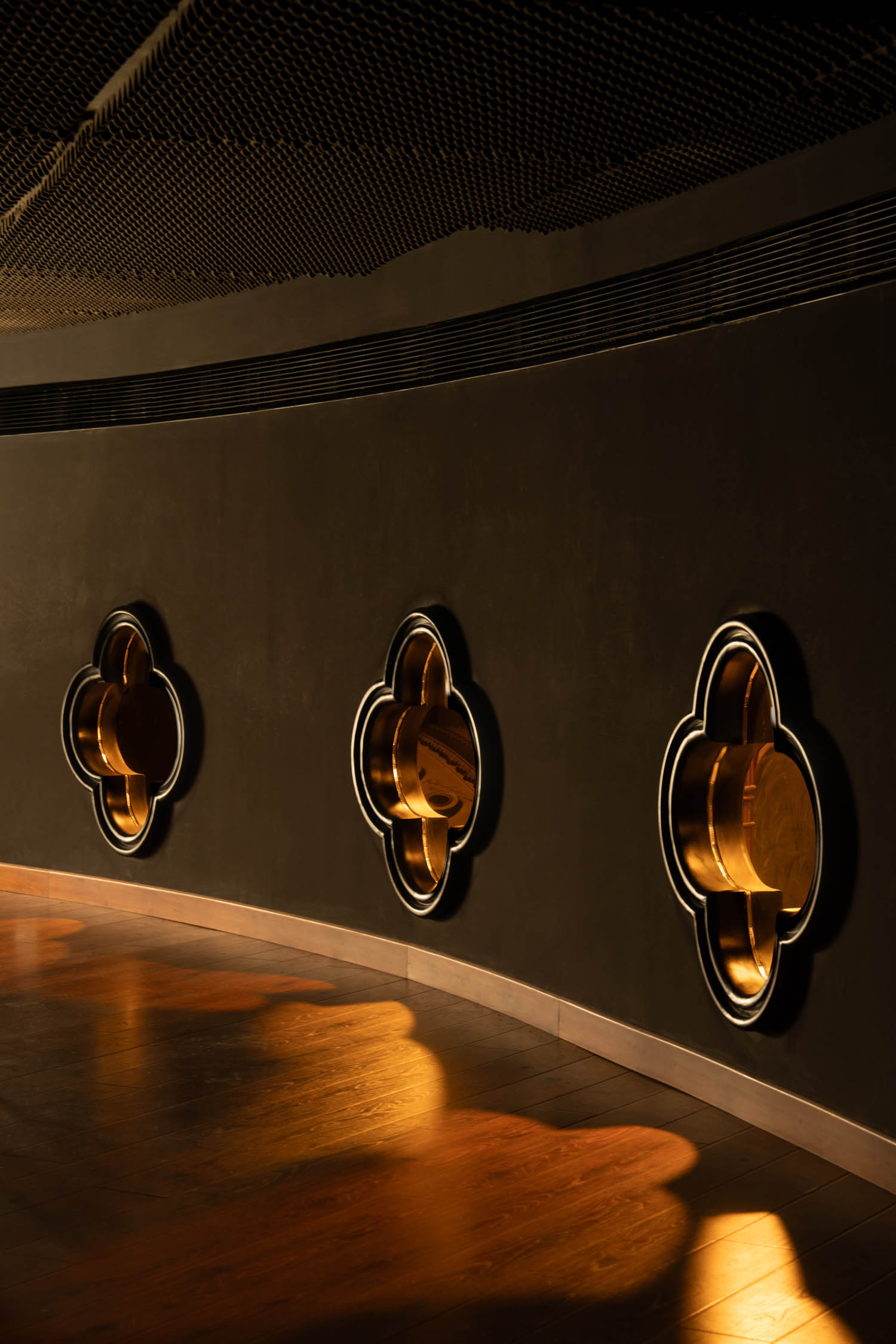 A curved dark wall features three gold clover-shaped fixtures with soft lighting casting warm reflections on the wooden floor.