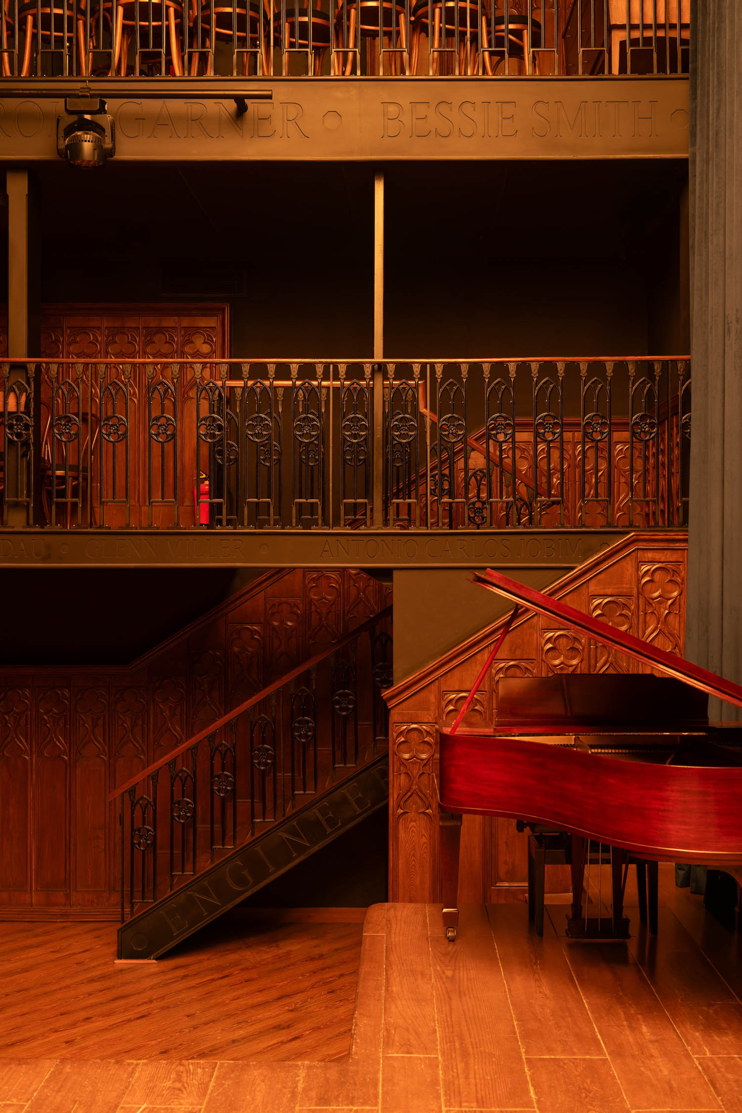 A red grand piano sits on a wooden stage beside an ornate balcony and staircase in a dimly lit theater.