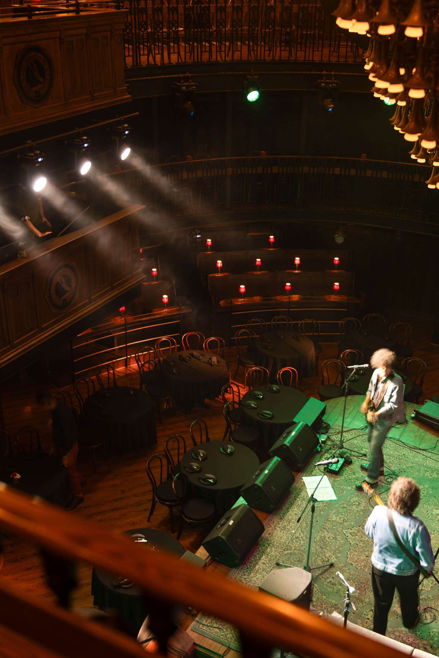 Two musicians perform on a dimly lit stage in a vintage-style theater with empty tables and chairs arranged on the main floor.