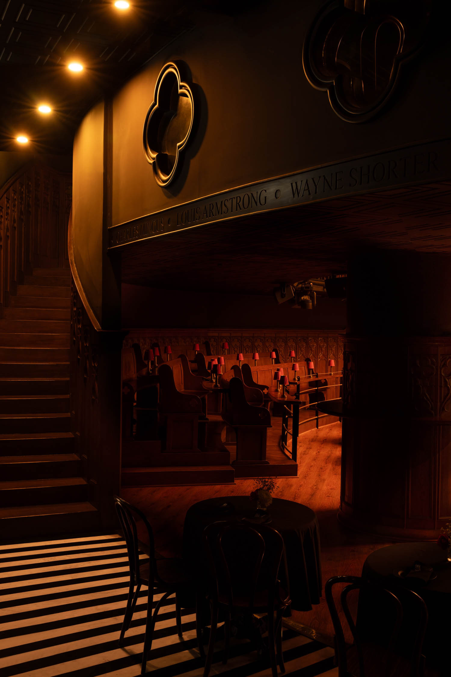 Dimly lit jazz club interior with empty tables and chairs, staircase on the left, and spotlights on the ceiling. Names of musicians carved above, including Louis Armstrong and Wayne Shorter.