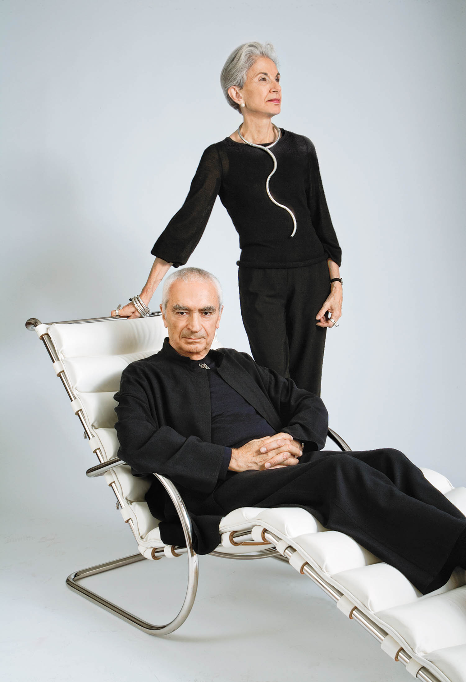 An older man in black sits on a modern white lounge chair, while an older woman in black stands behind him, one hand resting on the chair, both looking off camera.