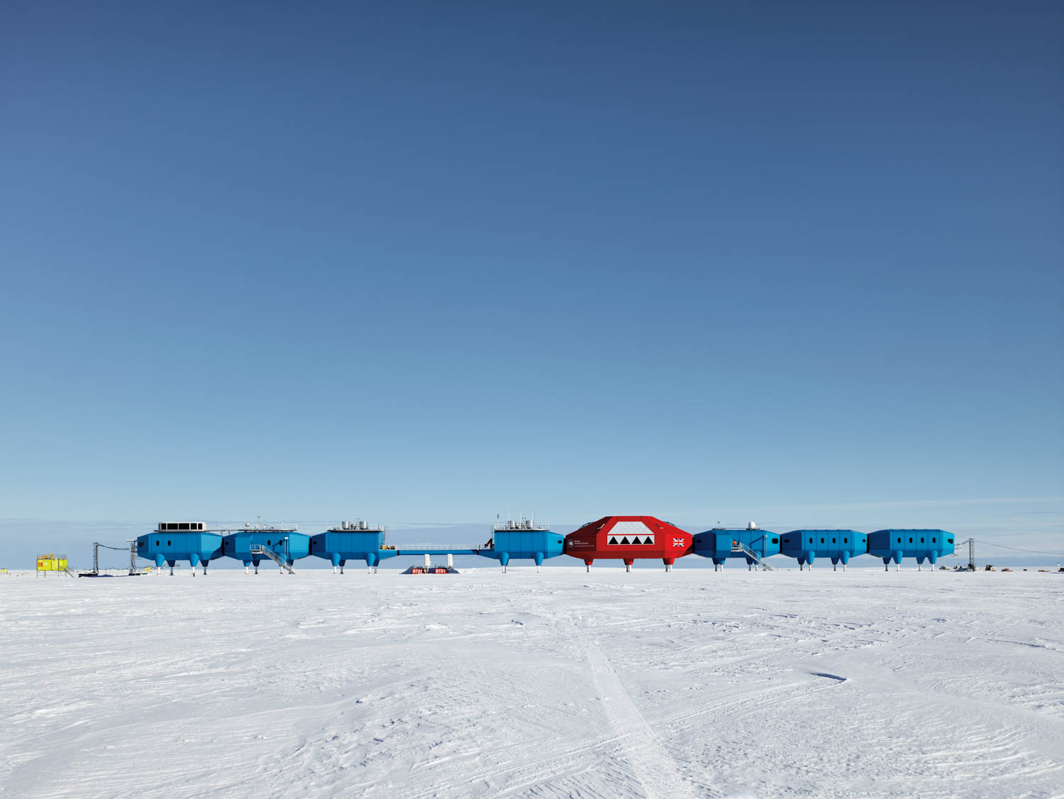 A row of blue and one red interconnected research modules stands on a snowy, flat landscape under a clear blue sky.