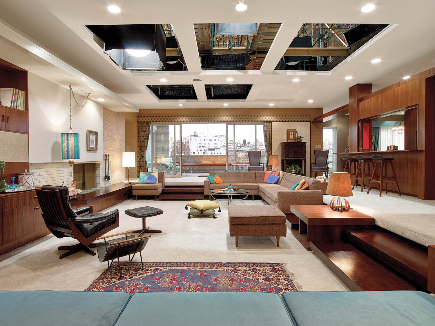 Spacious living room with mid-century modern furniture, large windows, sunken seating area, and ceiling panels revealing studio lights above.