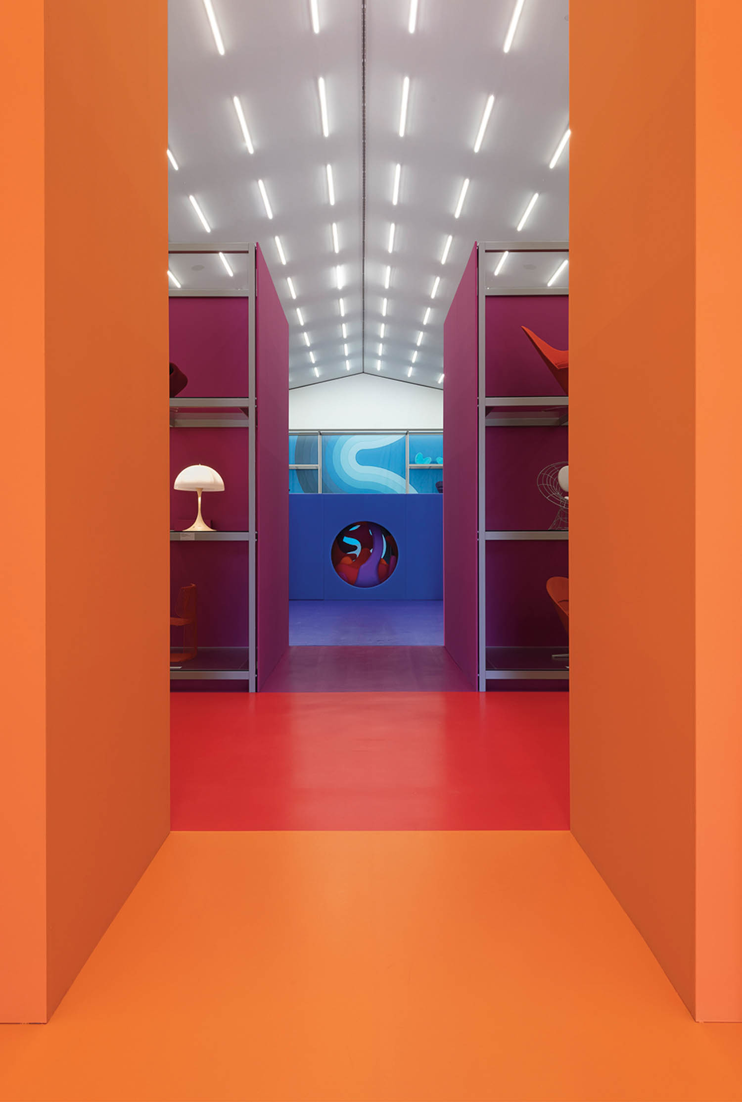 A brightly lit hallway with orange walls, red floor, purple shelves displaying objects, and a blue wall with a circular window in the background.