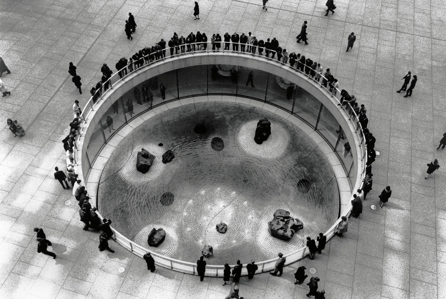 A large group of people stand around the edge of a circular water feature with rocks in a public plaza, seen from above.