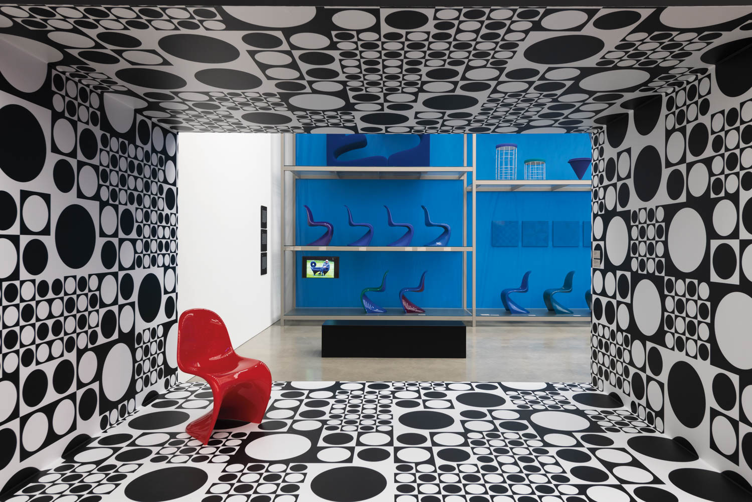 A room with black and white circular patterns, a single red chair, and a view into a blue-walled space with colorful chairs displayed on shelves.