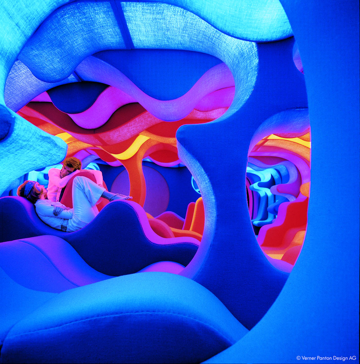 Two people sit in a vibrant, futuristic room filled with curving blue, pink, and purple sculpted forms and colorful ambient lighting.