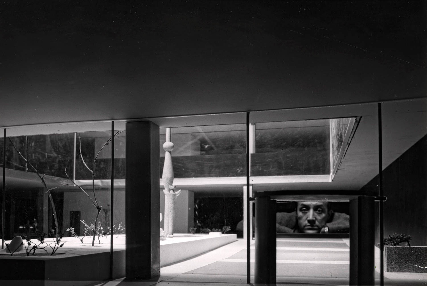 Black and white photo of a modernist architectural model with a man’s face visible through a window in the background.