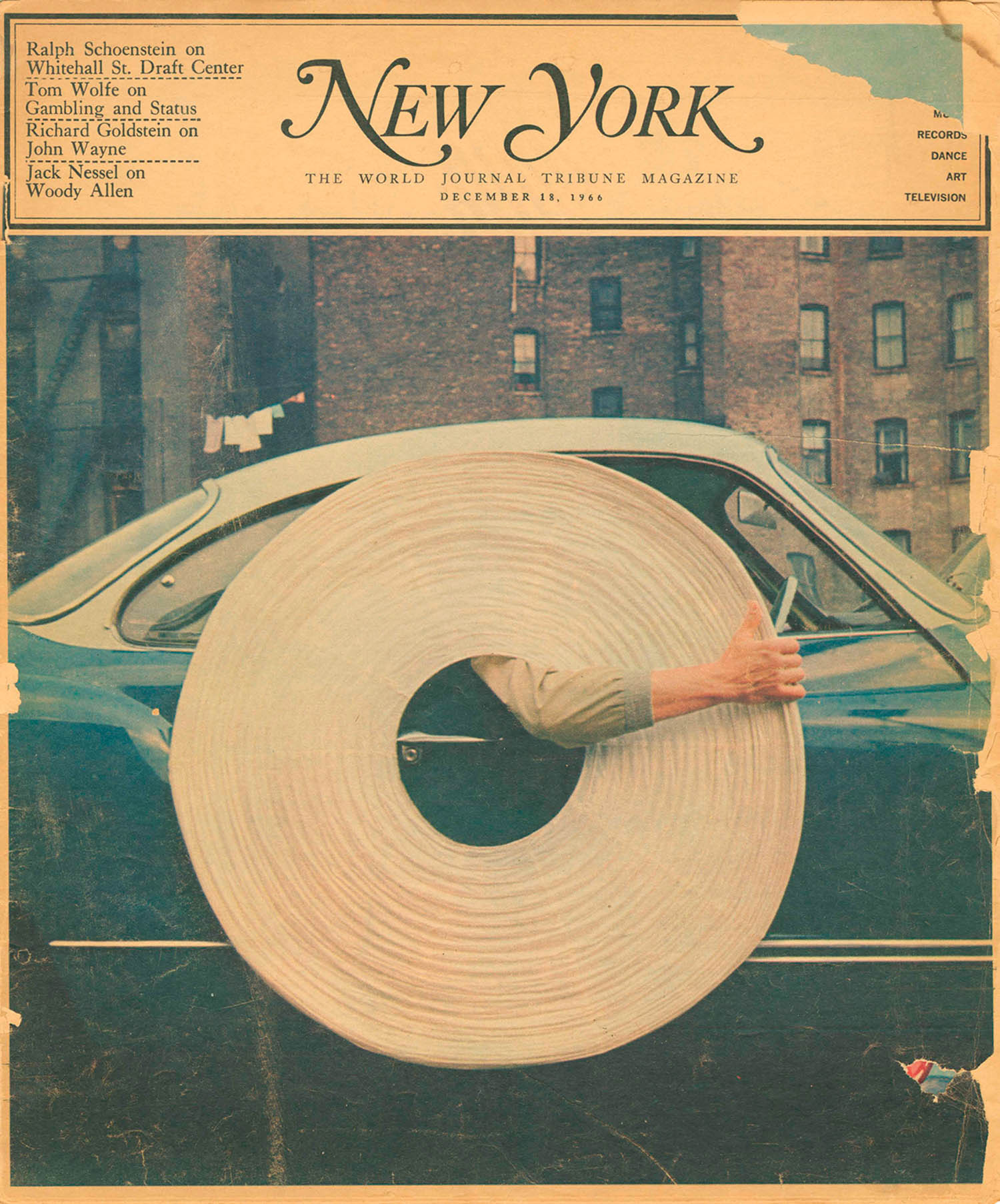 A person holds a giant roll of paper in front of a green car on a city street. The person’s face is not visible, only their arms and legs are seen behind the roll.