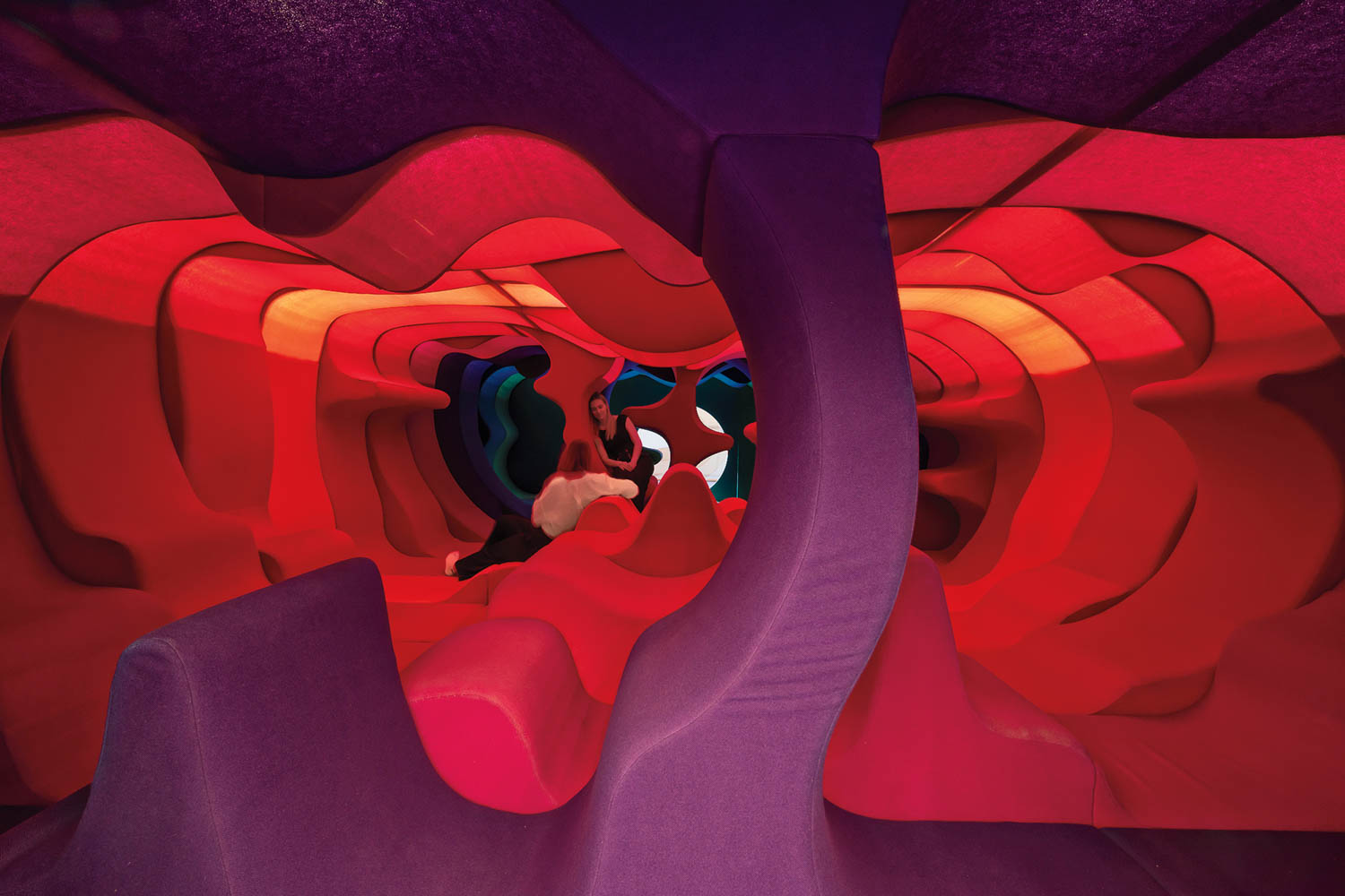 A person sits inside an abstract, colorful, cave-like structure with layered red, purple, and orange shapes forming the walls and ceiling.