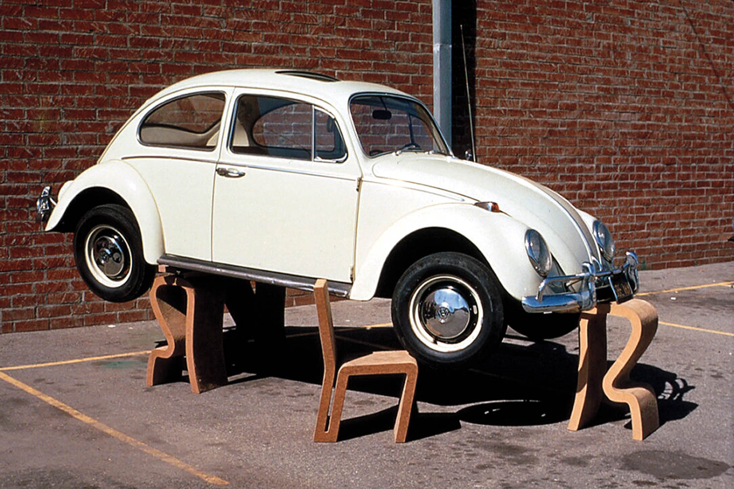 A white vintage Volkswagen Beetle is balanced on three wooden chairs in a parking lot next to a brick wall.