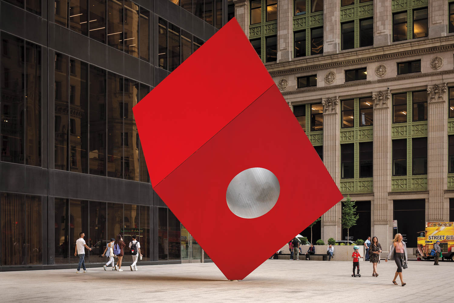 Large red cube sculpture with a silver circle on one face stands tilted in a city plaza as people walk nearby.
