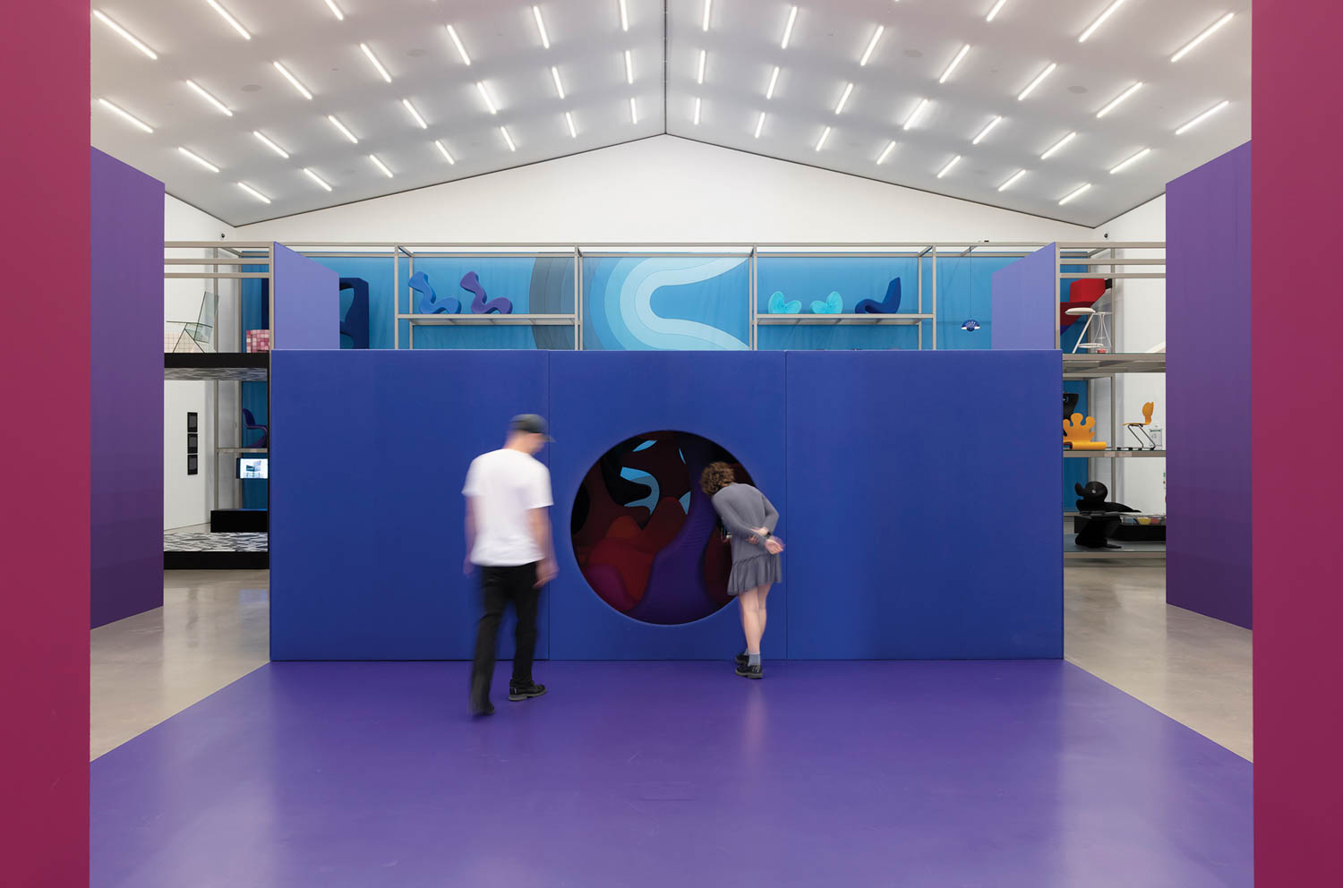 Two people stand in front of a circular entrance in a blue wall inside a brightly lit modern gallery with abstract art displays.