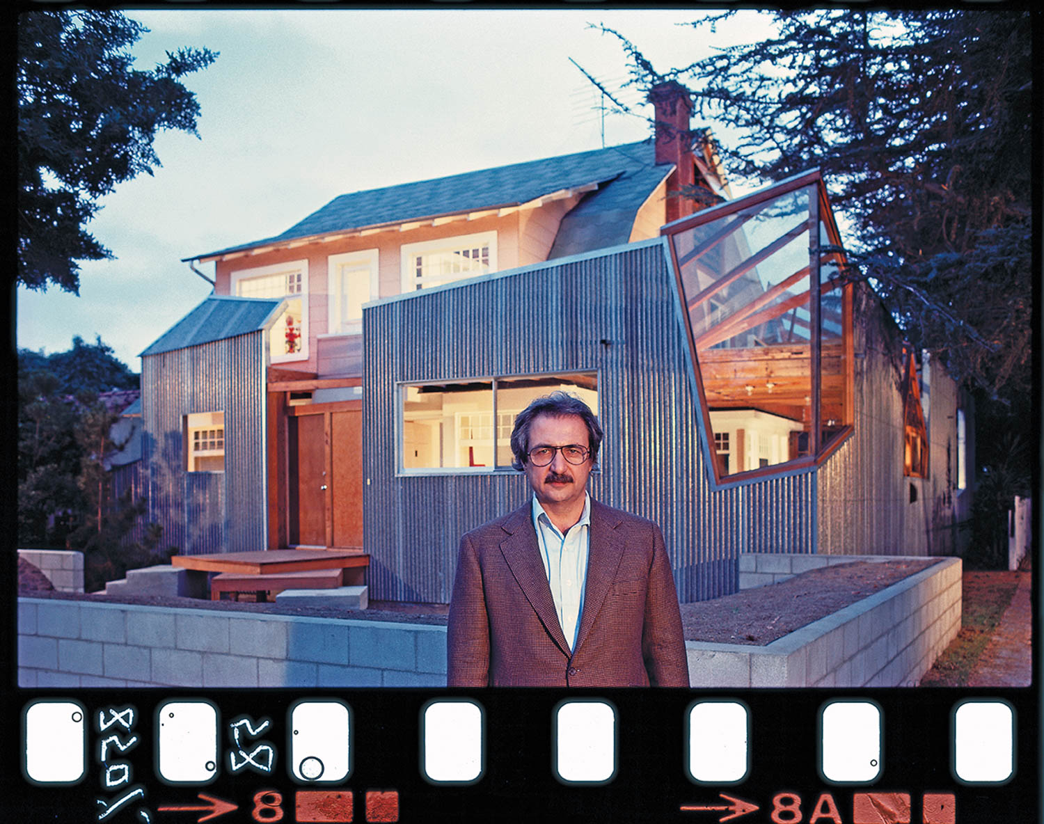 A man in a suit stands in front of a modern house with metal siding, large windows, and visible architectural modifications. The image includes a film strip border at the bottom.