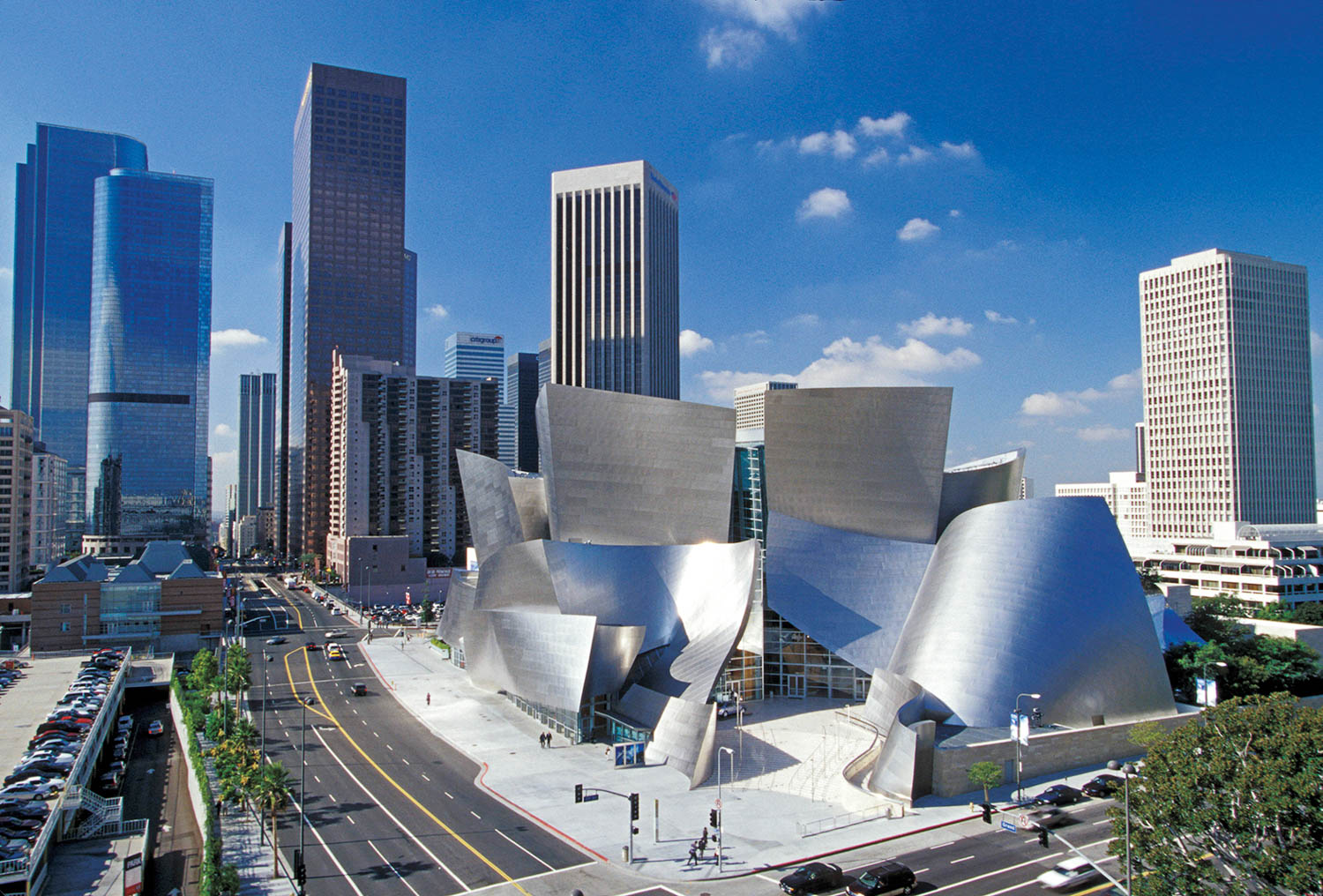 Walt Disney Concert Hall in downtown Los Angeles, featuring stainless steel curves, with surrounding skyscrapers and city streets under a blue sky.