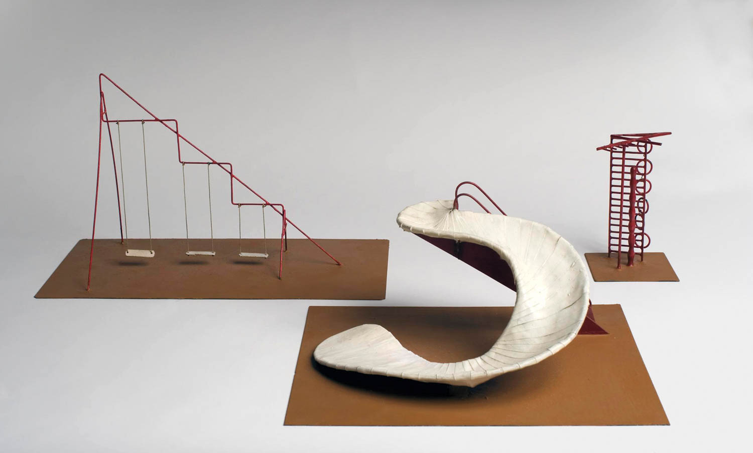 Three minimalist playground models: a red swing set, a red climbing structure with spiral steps, and a large white abstract slide, all set on brown rectangular bases against a plain background.