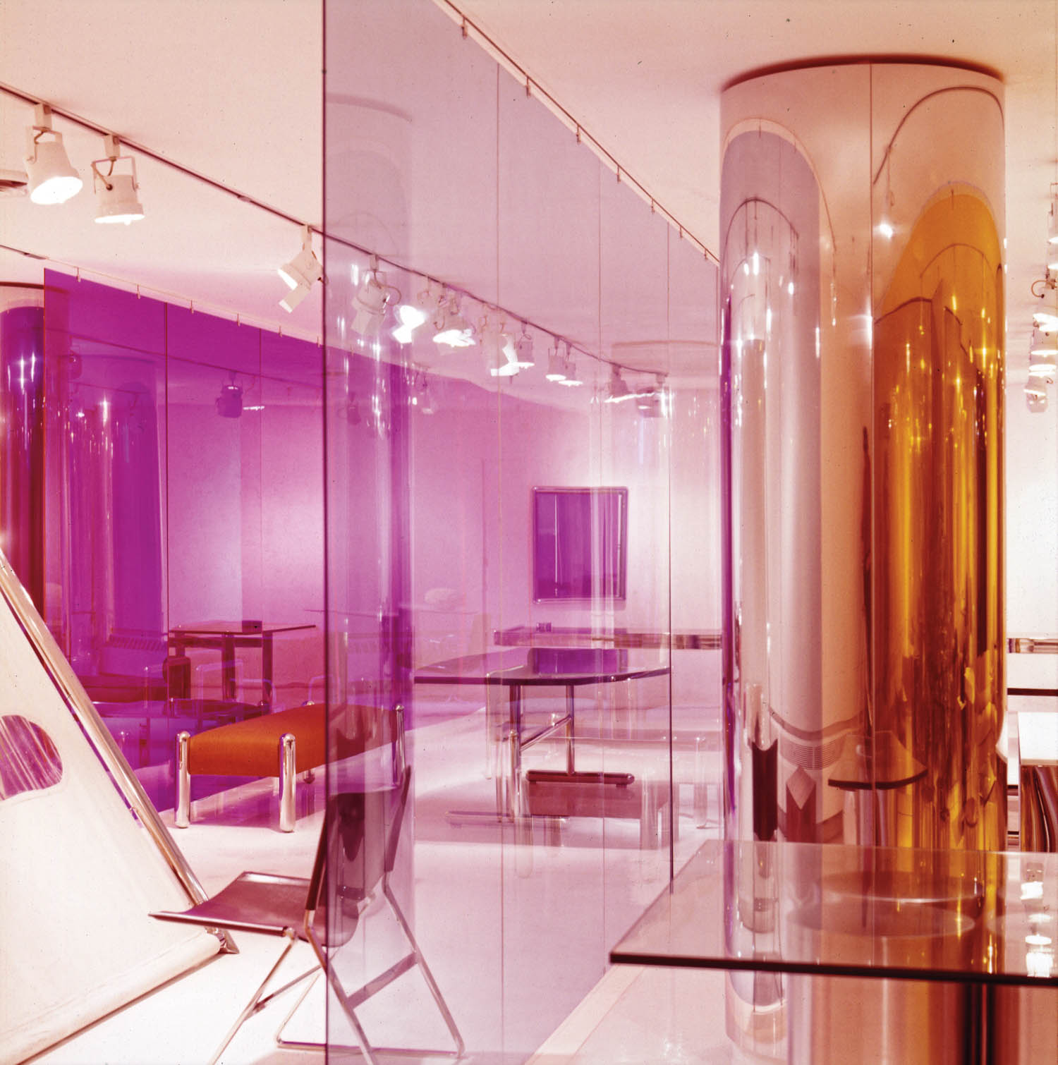 Modern interior with transparent purple and orange acrylic panels, reflective columns, minimal furniture, and bright ceiling lights. The space has a futuristic, open layout.