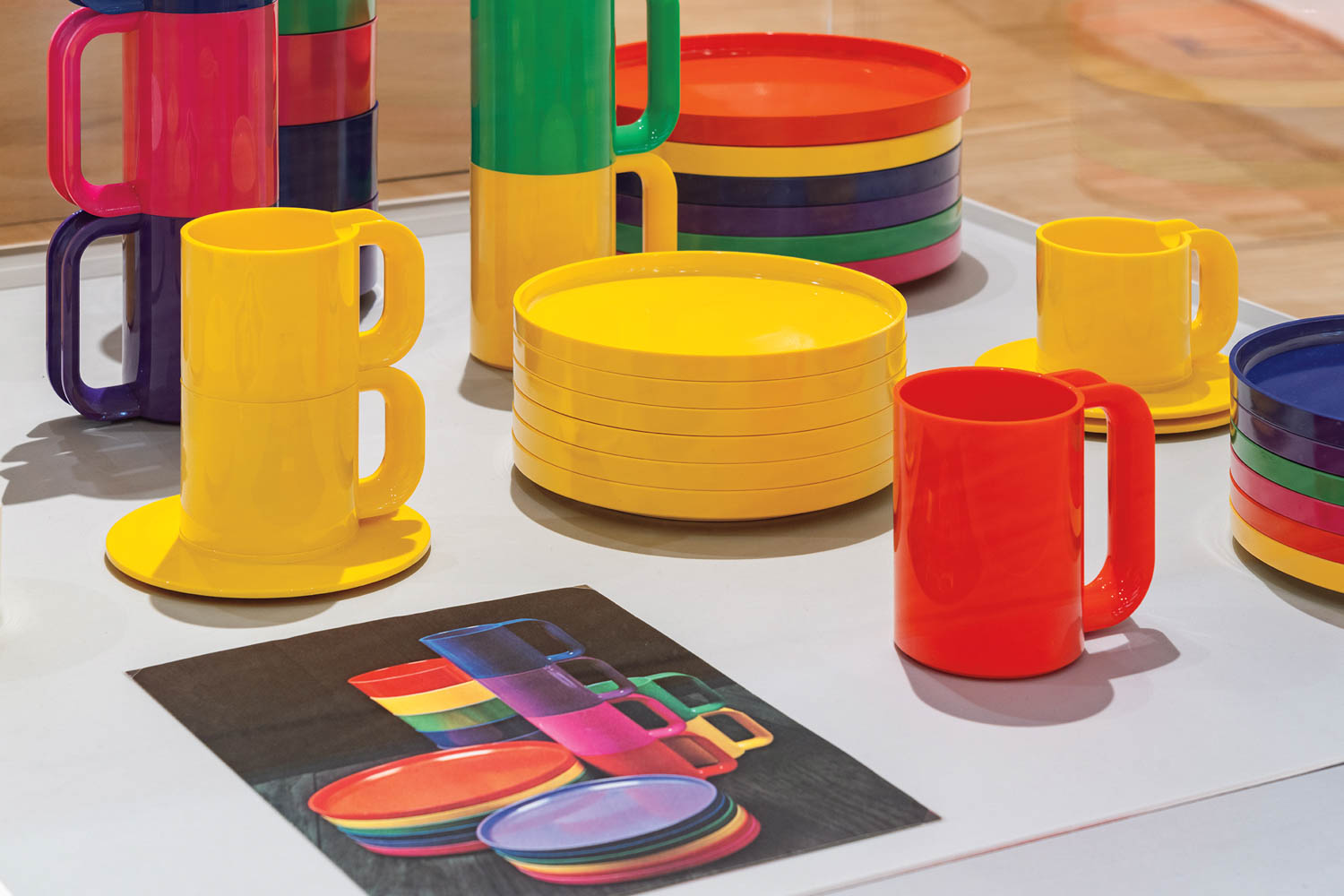 Colorful plastic cups, bowls, and saucers are neatly arranged on a table next to a drawing showing similar tableware in various bright colors.