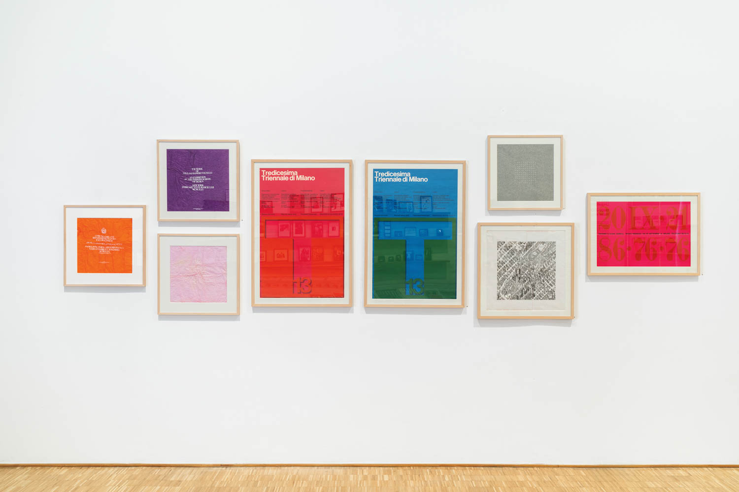 Eight framed posters and prints with colorful geometric and text designs are displayed in a row on a white gallery wall above a wooden floor.