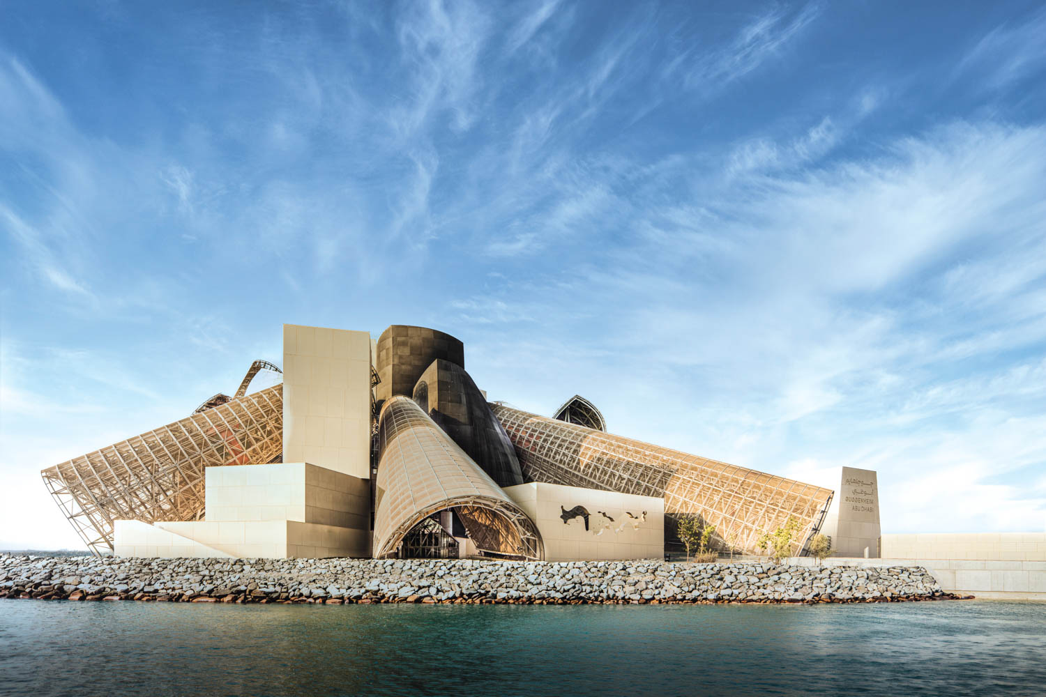 A modern building with angular, overlapping structures stands by the waterfront under a blue sky with wispy clouds.