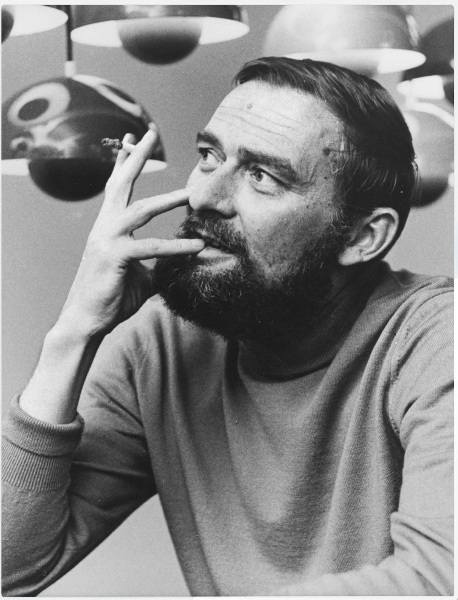 A bearded man in a turtleneck rests his hand on his face, looking upward thoughtfully, with spherical lamps in the background.