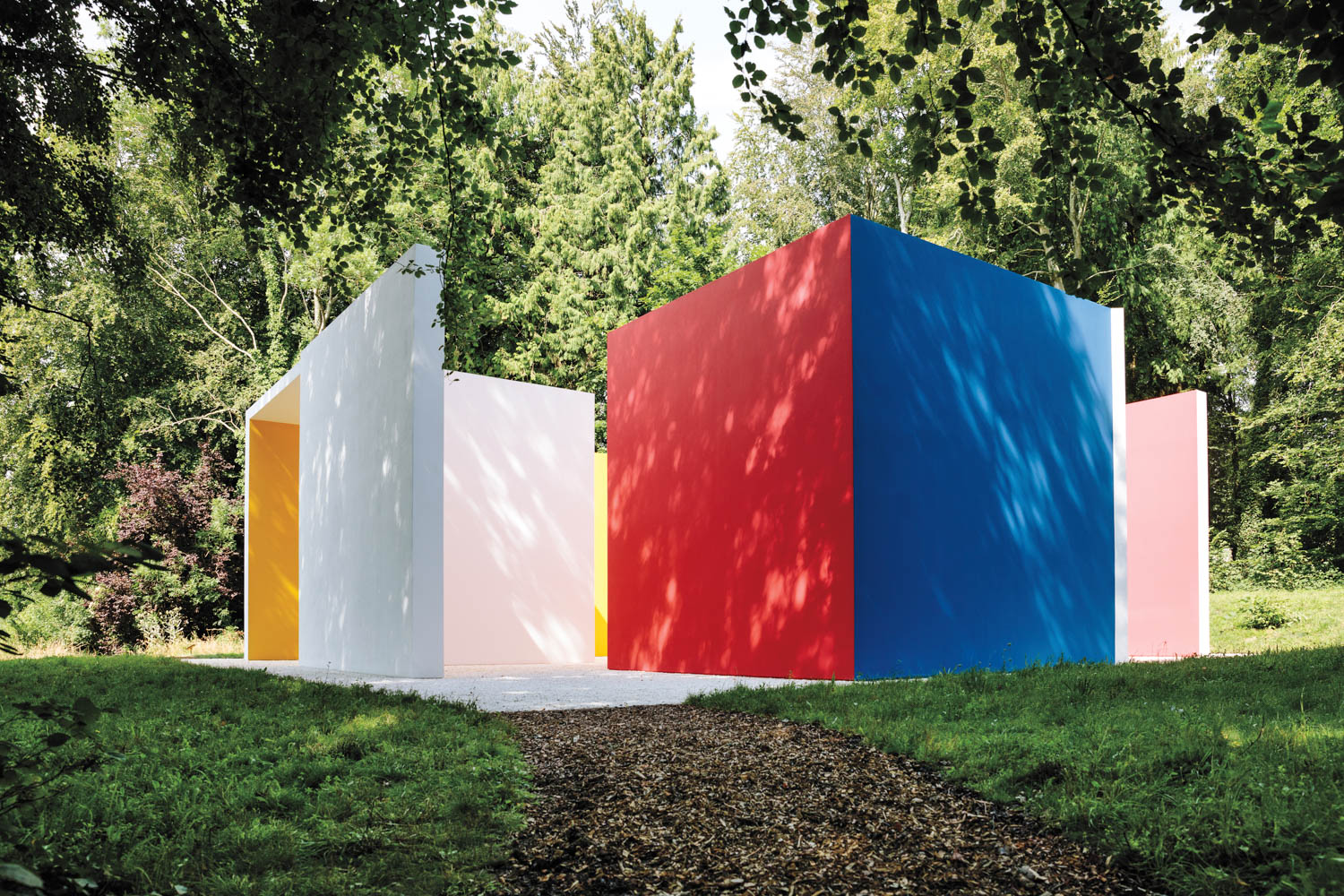 Colorful geometric outdoor structure with white, red, yellow, pink, and blue walls set on grass, surrounded by trees on a sunny day.