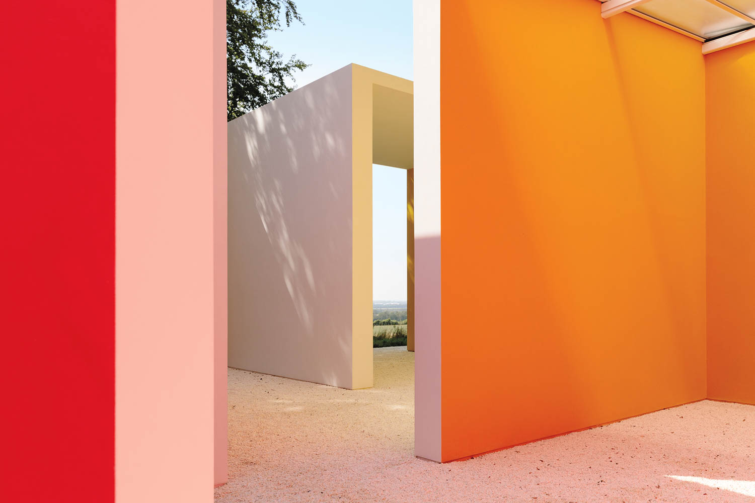Colorful geometric walls in shades of red, pink, orange, and white stand outdoors on a gravel surface under a clear blue sky.