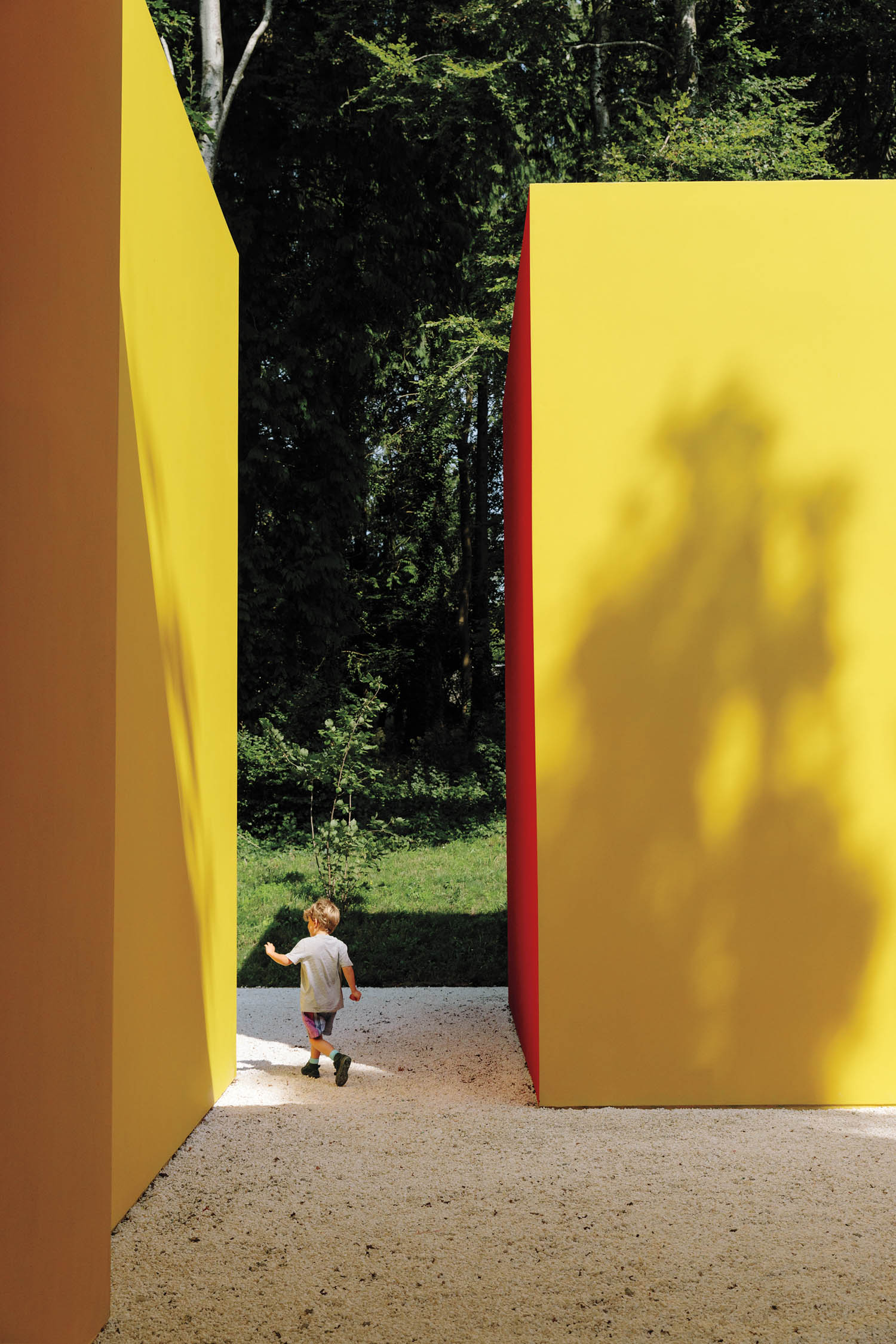A young child walks between large yellow and orange geometric structures outdoors, with trees and grass visible in the background.