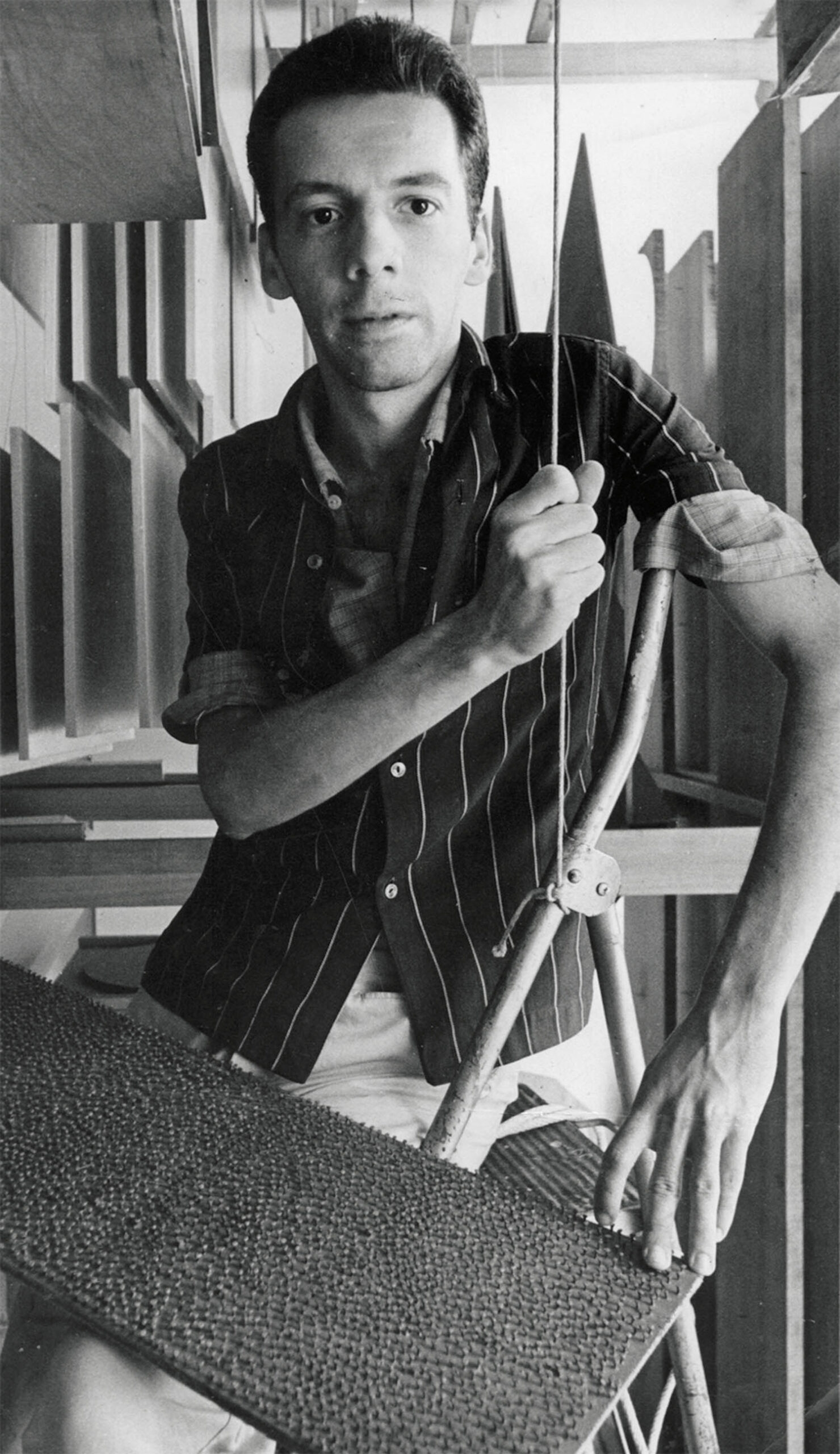 A man in a striped shirt sits on a chair, holding a thin vertical rod, with one hand resting on a textured board in a room filled with geometric objects.