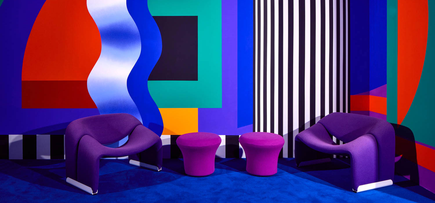 Two modern purple chairs and two round stools sit in a room with a bold, colorful geometric and striped wall design and blue carpet.