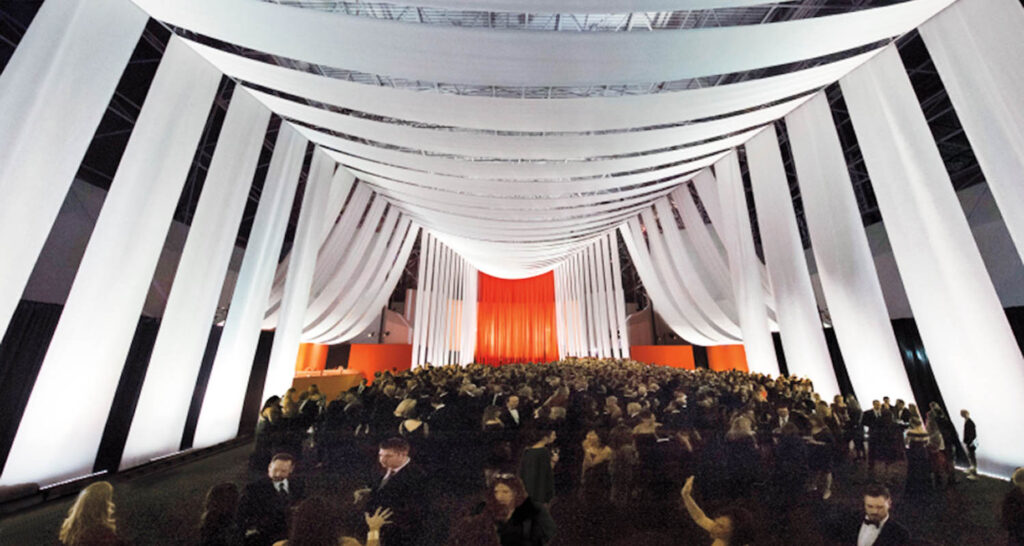 A large crowd in formal attire gathers under dramatic white drapes hanging from the ceiling in a spacious, elegantly lit venue.