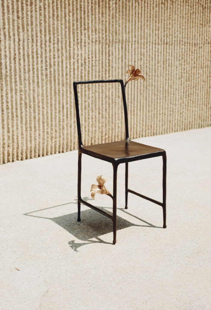 A metal chair with a minimalist design features two sculpted lilies on the backrest and leg, positioned on a sunlit concrete surface against a textured wall.