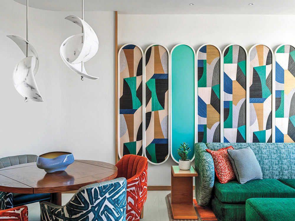 Modern living room with colorful geometric wall panels, green sofa, patterned chairs around a wooden table, white pendant lights, and a small plant on a side table.