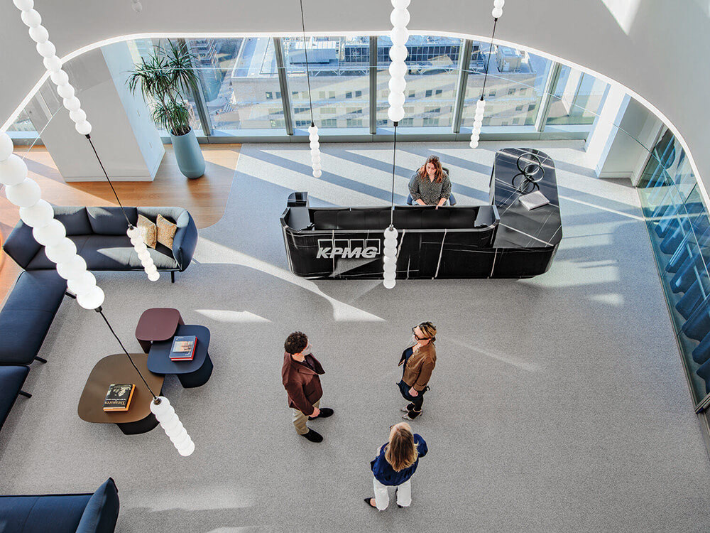 Three people stand talking near a curved black reception desk in a modern, bright office lobby with large windows and hanging lights.